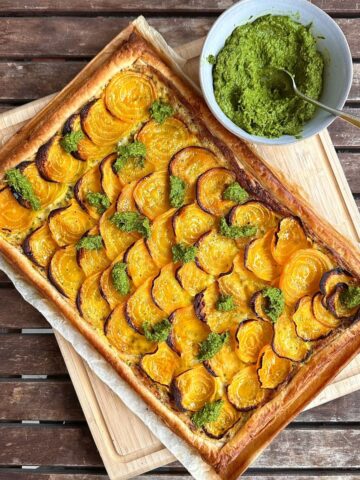 A beetroot tart garnished with beetroot leaf pesto, cut into 8 slices, sitting on parchment paper and a wooden chopping board. A bowl of beetroot leaf pesto sits next to the tart.
