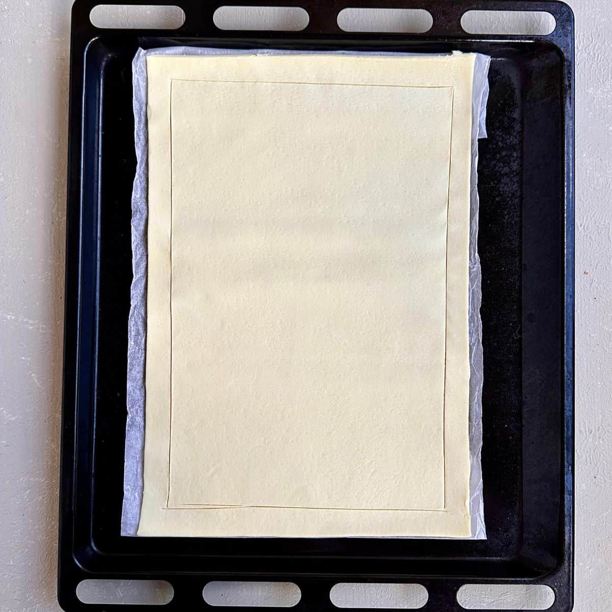 A sheet of puff pastry scored with a knife around the edges, sat upon parchment paper and a baking tray. Foe a beetroot tart recipe.