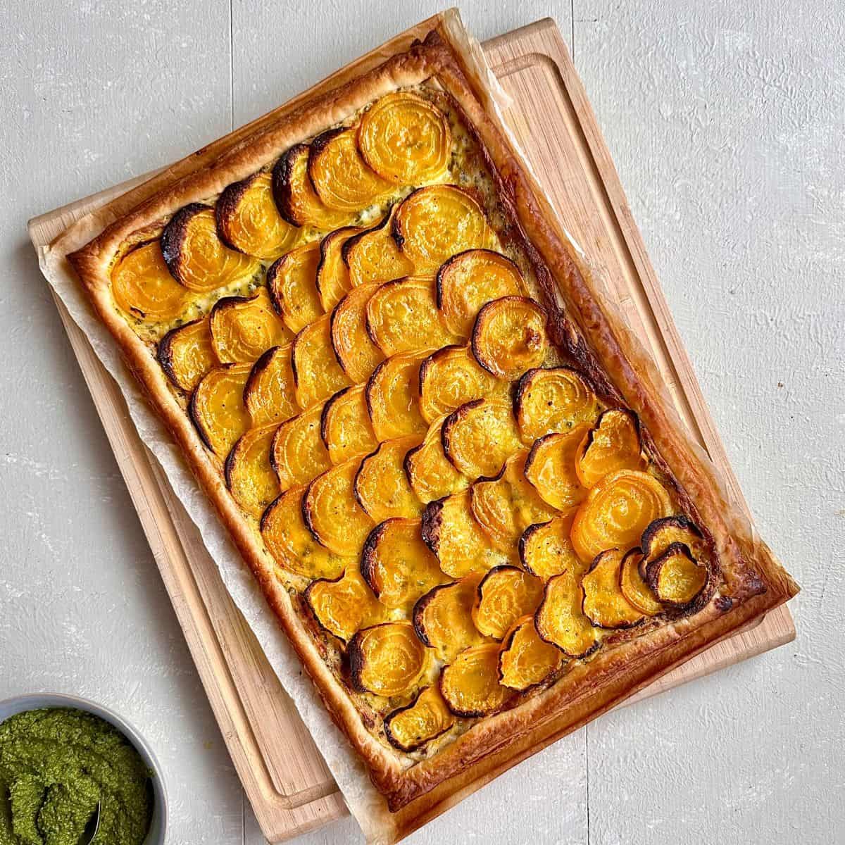 A freshly baked beetroot tart sat on parchment paper and a wooden chopping board. A bowl of beetroot leaf pesto sits next to the chopping board.