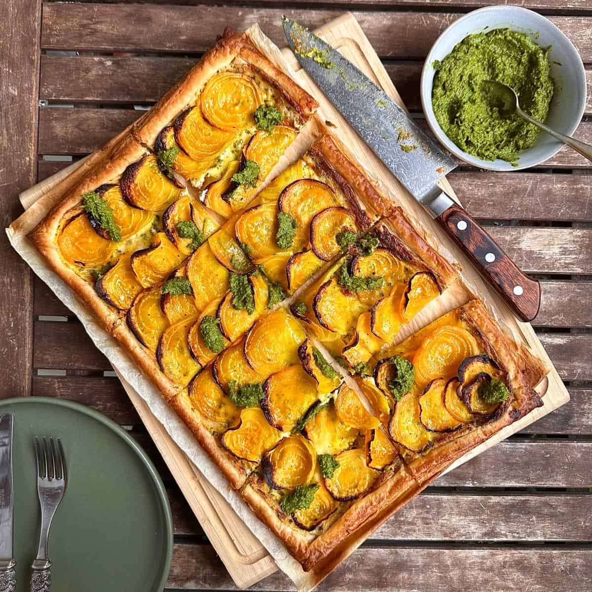 A beetroot tart with mascarpone cut into 8 slices, sitting on parchment paper on a wooden chopping board. A knife sits next to the tart.