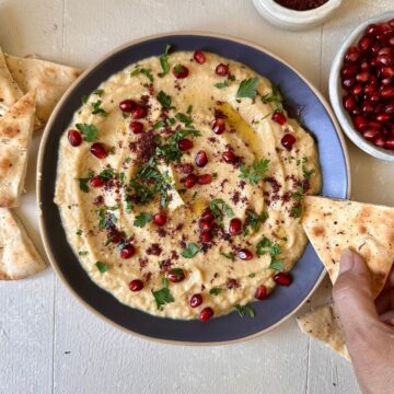 A navy blue plate containing courgette moutabbal garnished with sumac, parsley and pomegranate. A hand holds pita bread over the plate.
