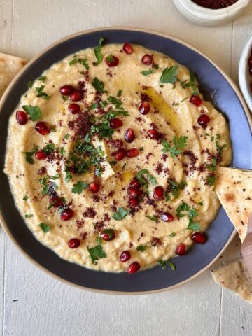 A navy blue plate containing courgette moutabbal garnished with sumac, parsley and pomegranate. A hand holds pita bread over the plate.