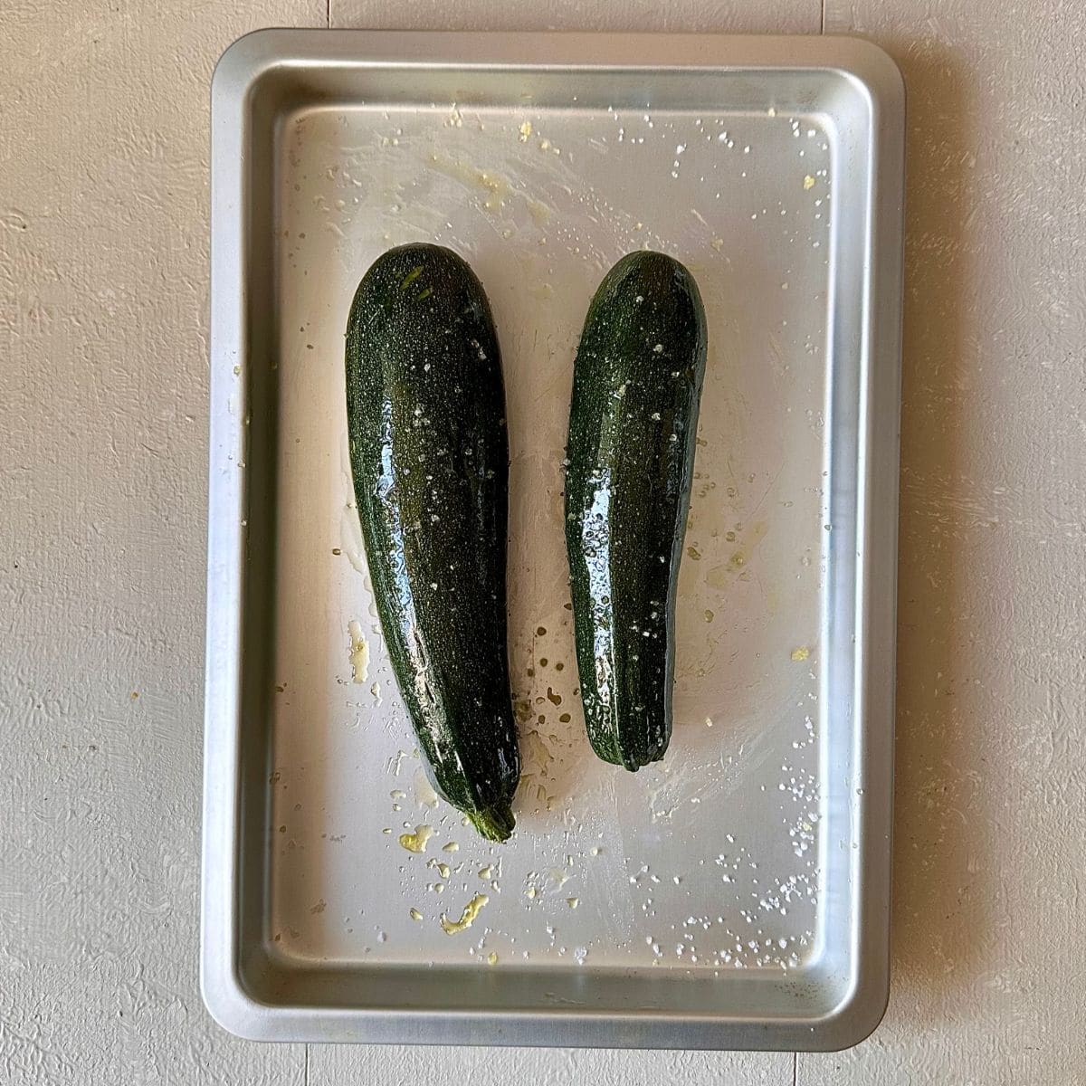 A baking tray containing two whole green courgettes coated in olive oil and sea salt ready for baking, for courgette moutabbal.