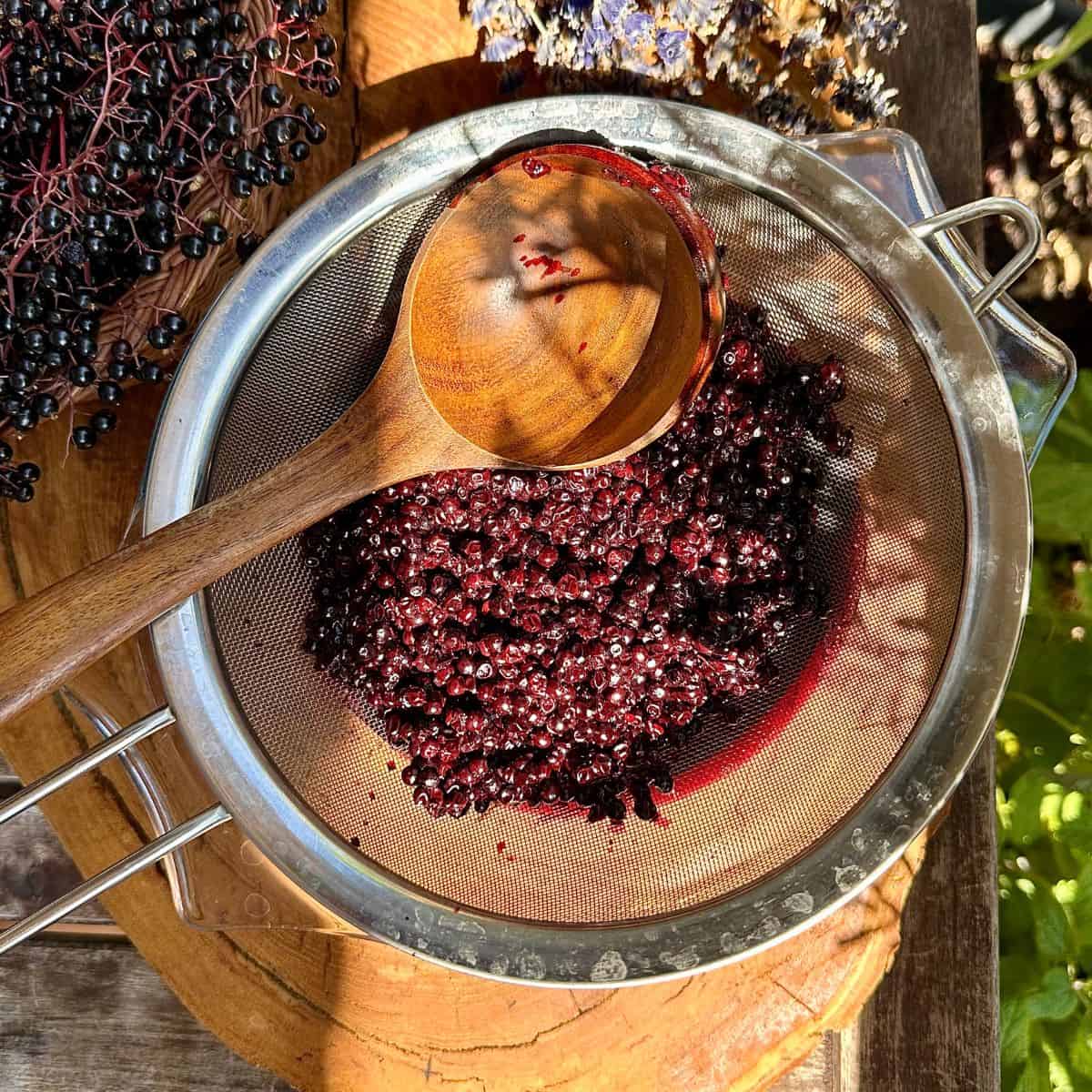 Elderberries in a sieve with a wooden ladle squeezing the elderberry juice into a glass dish. For elderberry lavender ice cream.