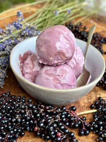 A dessert bowl containing three scoops of elderberry and lavender ice cream. The bowl is surrounded by elderberries on their stems and lavender.