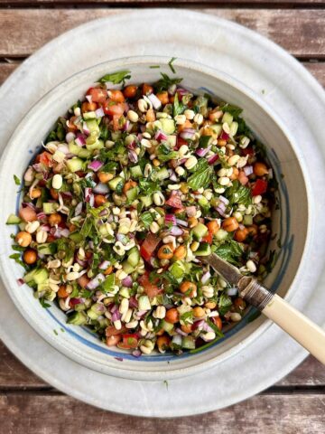 Bowl on a plate containing kachumber salad with kala chana and green mung bean sprouts. A spoon sits in the bowl.