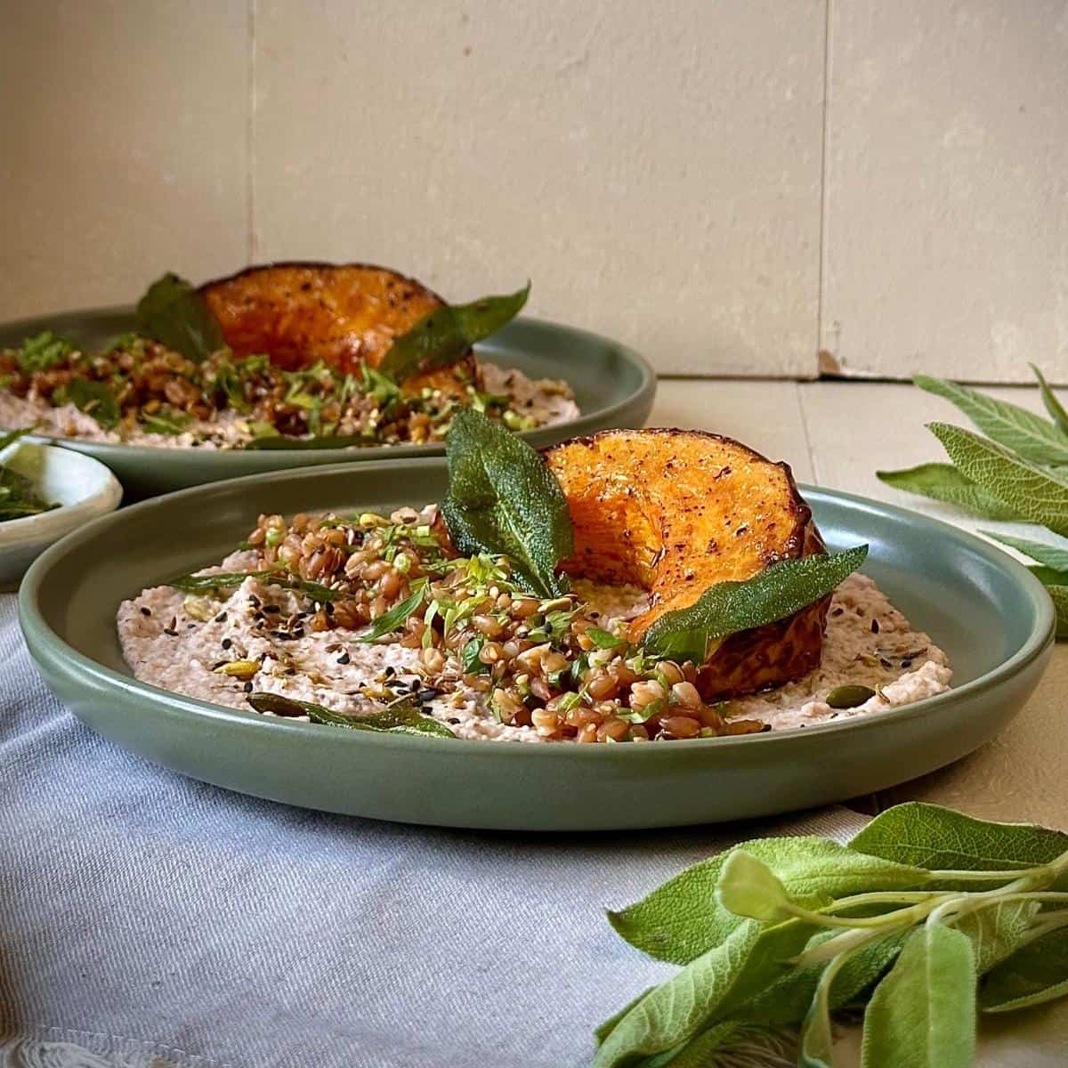 A green plate containing roasted pumpkin on a bed of black eyed pea hummus, garnished with sage leaves.