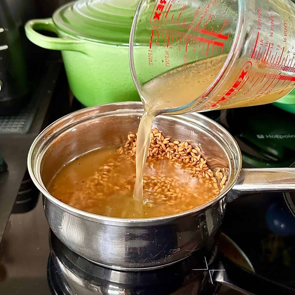 A steel saucepan containing spelt in vegetable stock. Vegetable stock is being poured into the pot from a measuring jug. For roasted pumpkin black eyed pea hummus.