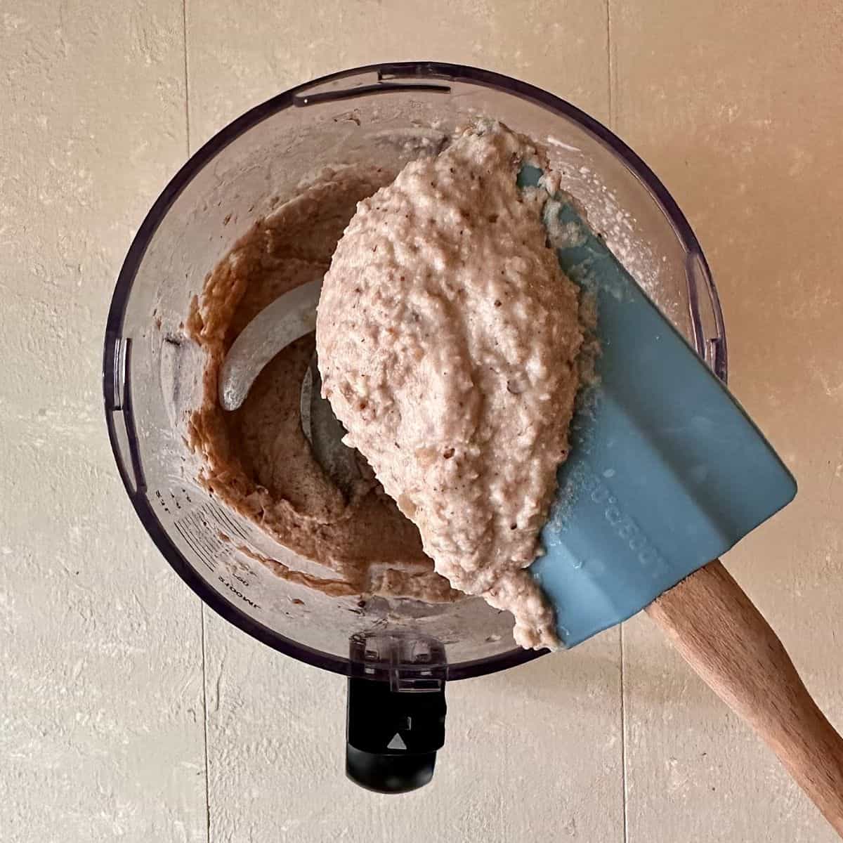 A blender jug containing black eyed pea hummus after blending for roasted pumpkin and black eyed pea hummus recipe. A spatular is held above the blender jug with the hummus on it.