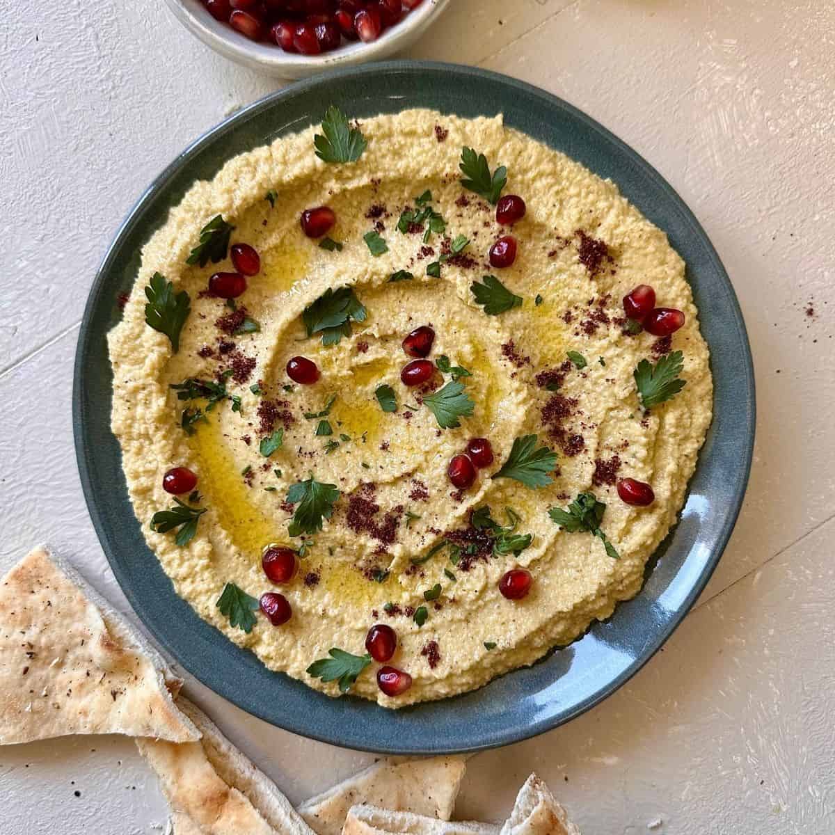 A blue ceramic plate spread with watermelon rind hummus, garnished with parsley and pomegranate seeds.