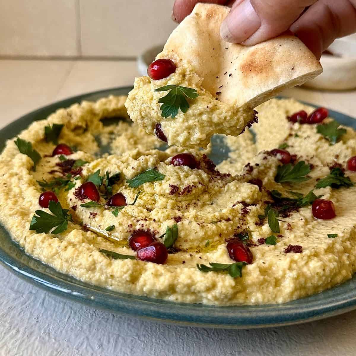 close up of a plate containing watermelon rind hummus, garnished with pomegranate fruit, parsley and sumac. A hand holds a piece of pita bread over the hummus with some of the hummus scooped onto it.