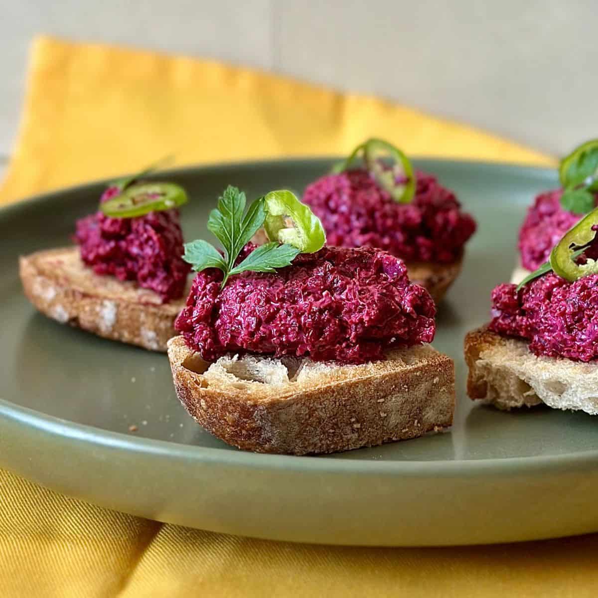 Beetroot aguachile left-over pulp mixed with cream cheese and spread onto small slices of sourdough bread on a plate. Garnished with a coriander leaf and slice of chilli.