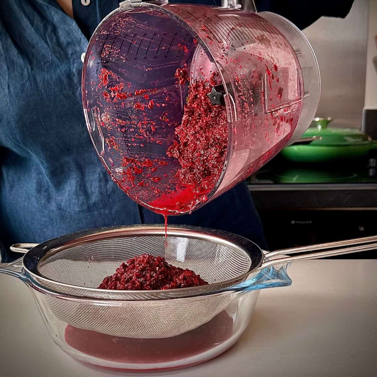 Roughly blended ingredients for beetroot aguachile being poured from a jug blender into a sieve. The juice is being collected in a glass bowl under the sieve.