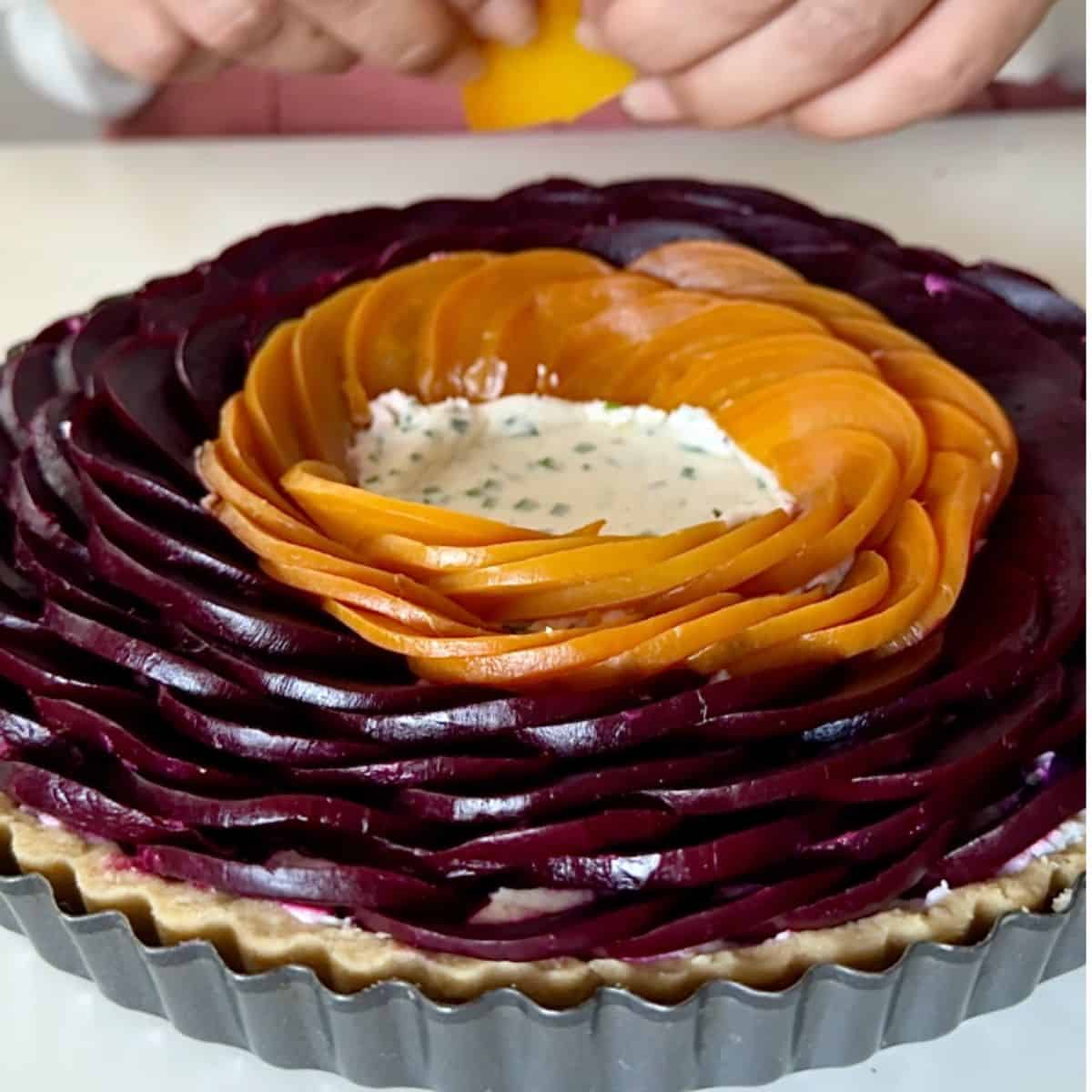 A red and golden beetroot tart in a fluted tart tin. Thin slices of beetroot are being laid in concentric circles on top of the whipped goats cheese filling. For a beetroot and goats cheese tart.