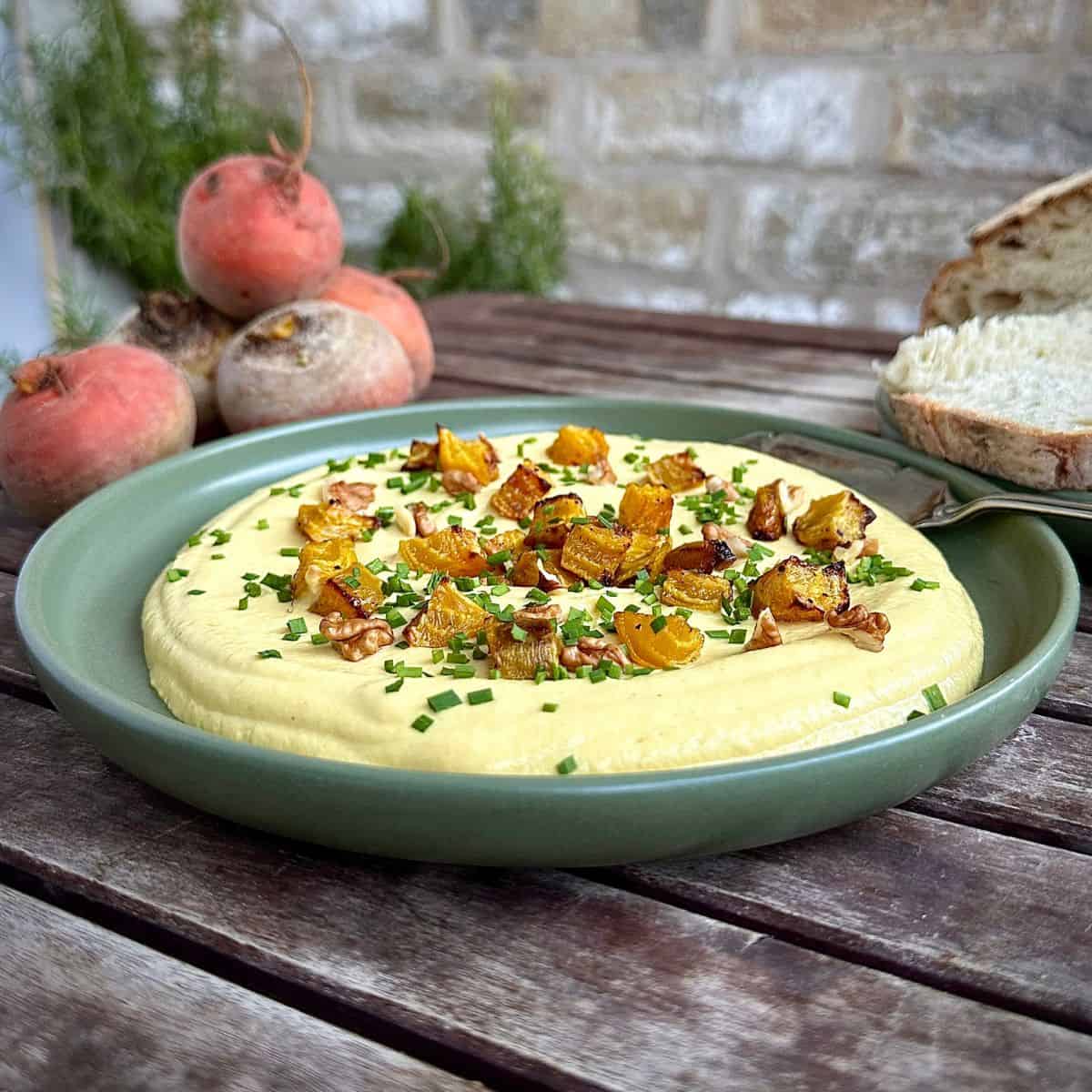 A green plate containing golden beetroot dip garnished with roasted golden beetroot and chives. Whole golden beetroot and bread sit in the background.