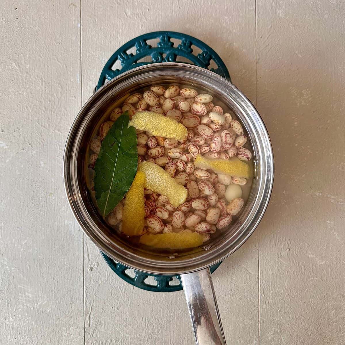 A steel saucepan containing borlotti beans in water with lemon peel, bay leaf and garlic. Ready to boil for borlotti bean salad recipe.