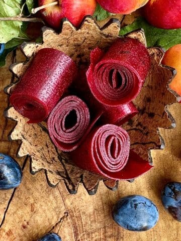 Four rolls of damson and crap apple fruit leather in a carved coconut cup. Surrounded by whole damsons and crap apples for decoration.