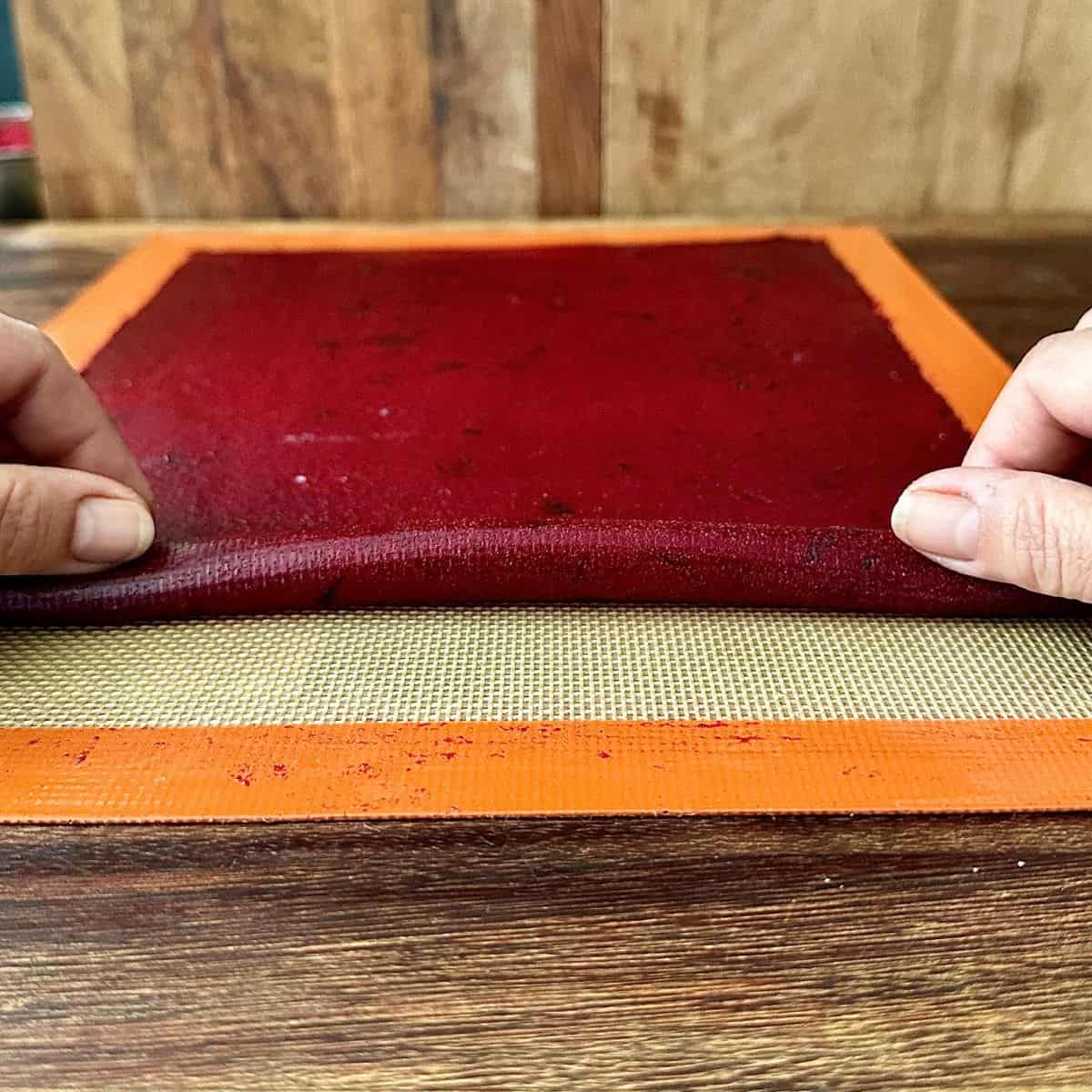 A crab apple and damson fruit leather sheet on a silicone mat being rolled into a log after cooking.