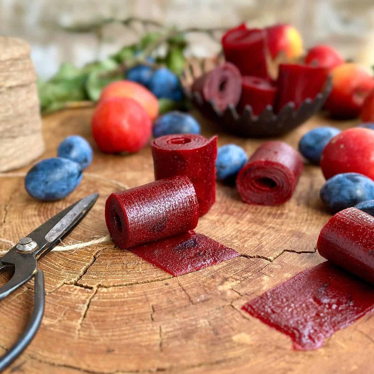 A rustic wooden platter with cut rolls of crab apple and damson fruit leather. Whole damsons and crab apples lay around for decoration.