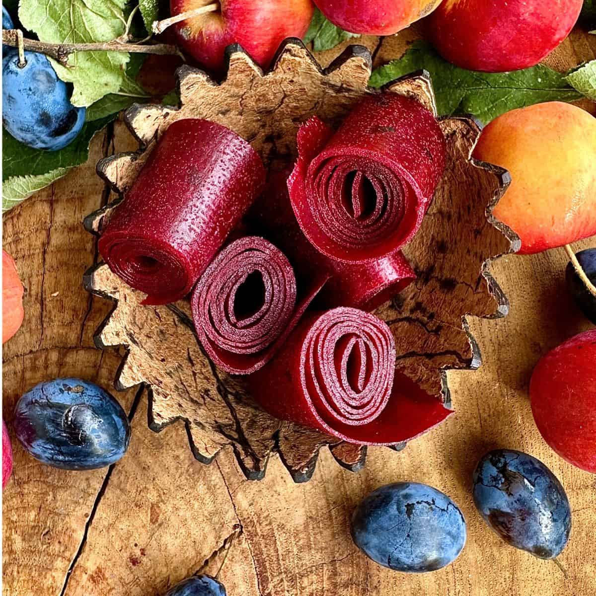 Four rolls of damson and crap apple fruit leather in a carved coconut cup. Surrounded by whole damsons and crap apples for decoration.