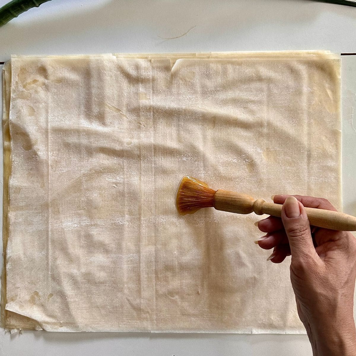 A hand holding a pastry brush brushes melted butter onto sheets of filo pastry. For a leek tart tatin recipe.