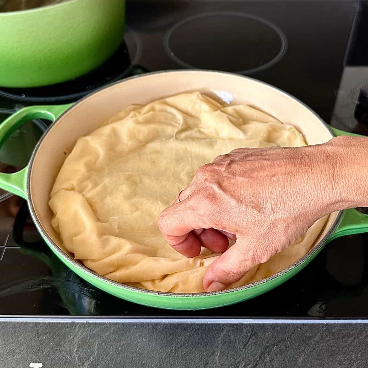 A large green oven-proof dish atop a stove top containing leeks covered with filo pastry. A hand tucks the pastry inside the dish. For a leek tart tatin recipe.