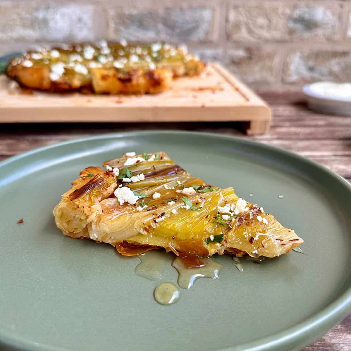 A slice of leek tarte tatin on a green plate, drizzled with honey. The rest of the tarte sits in the background, blurred.