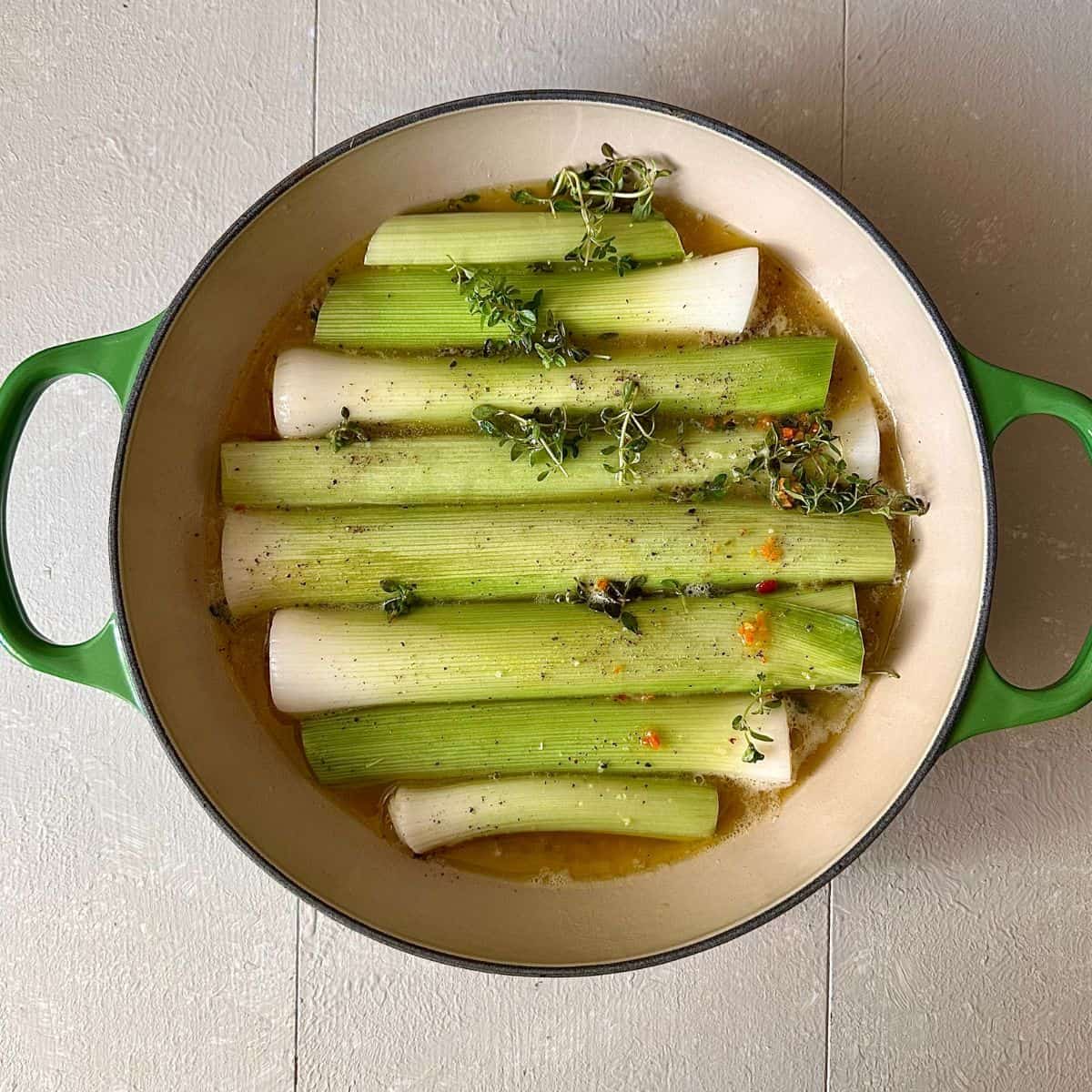 A large green oven-proof dish containing leeks, melted butter, thyme sprigs and vegetable stick, ready to cook for a leek tarte tatin.
