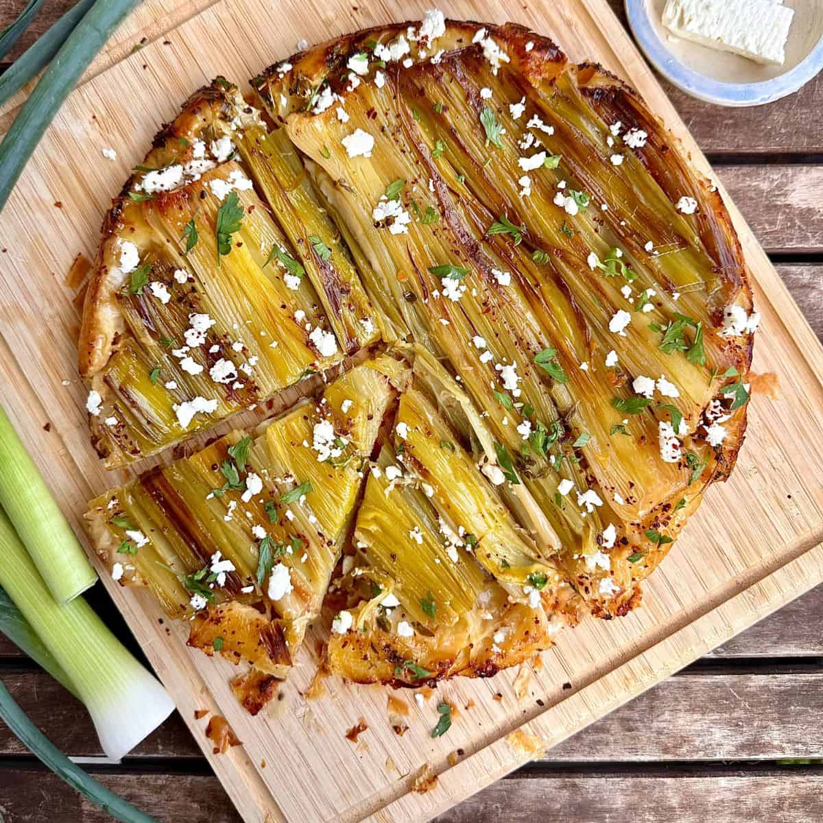 A leek tarte tatin sat on a wooded chopping board, with whole leeks sat beside the chopping board.