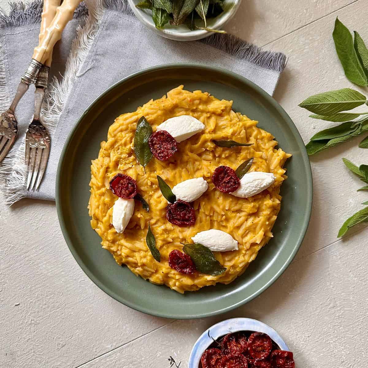 A green plate containing pumpkin orzo, garnished with soft cheese guenelle, fried sage leaves and dried cherry tomatoes.