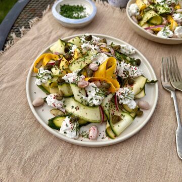 A plate containing ribboned courgette salad with ricotta cheese and borlotti beans and capers. garnished with rosemary sprigs and chives.