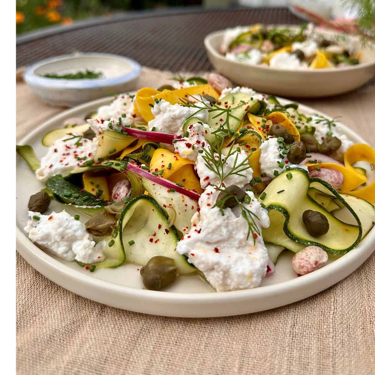 A white plate containing ribboned courgette salad with ricotta cheese, capers and borlotti beans.
