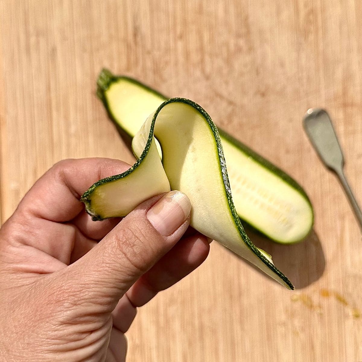 A hand holding a thin ribbon of green courgette close to the camera. For ribboned courgette salad.