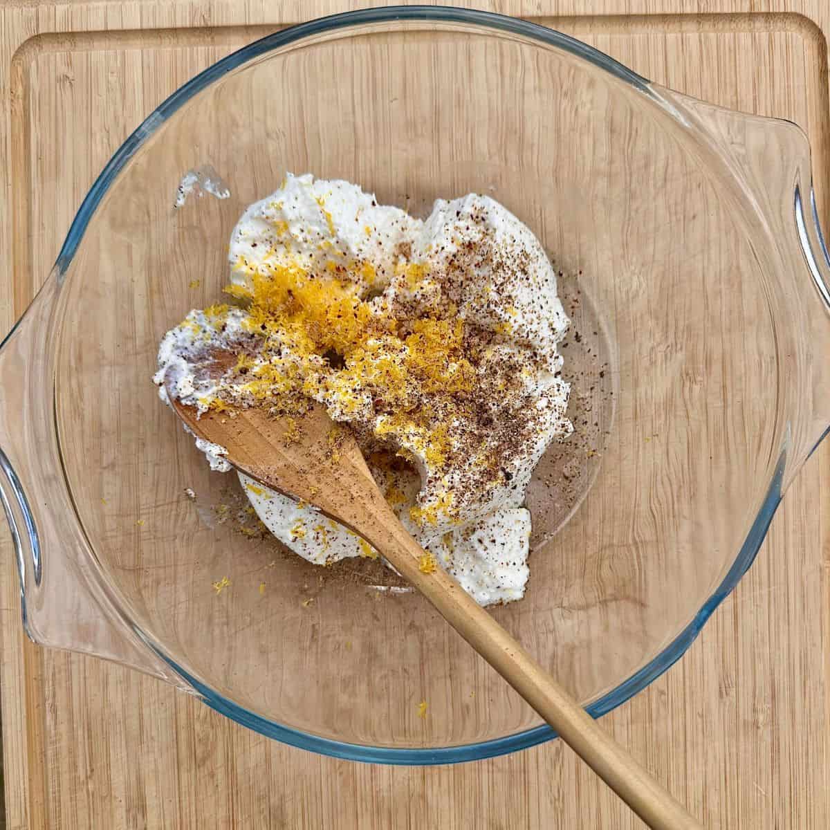 A glass bowl containing ricotta cheese, lemon zest, salt and pepper and gochujang, ready to mix together for ribboned courgette salad. A wooden spoon sits in the bowl.