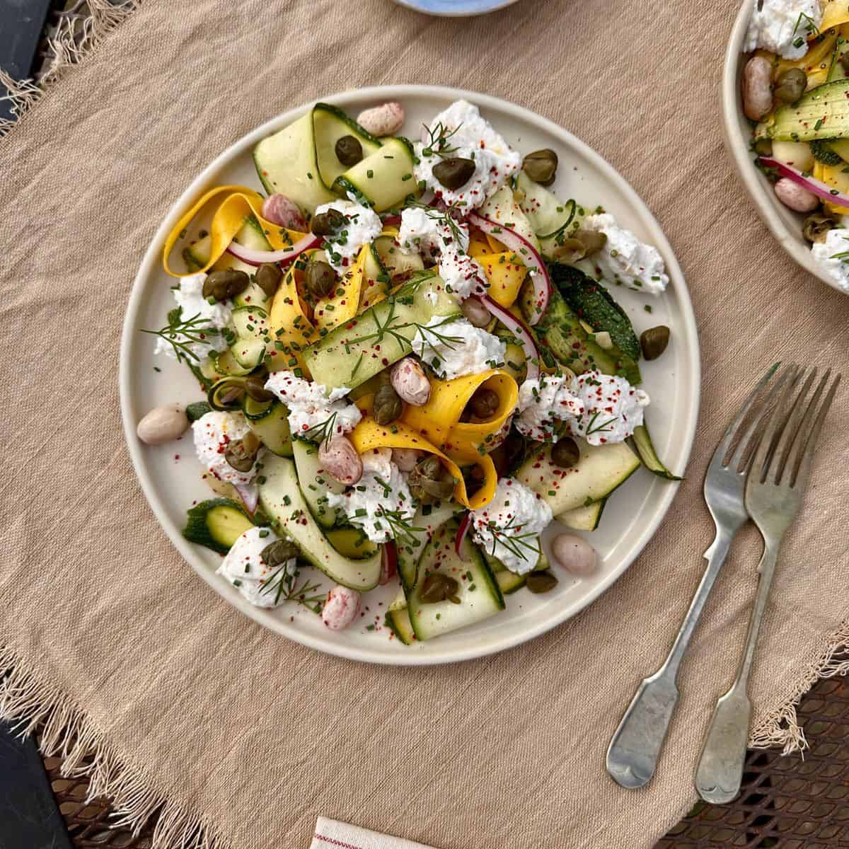A beige plate containing ribboned courgette salad with borlotti beans and ricotta, garnished with rosemary sprigs.