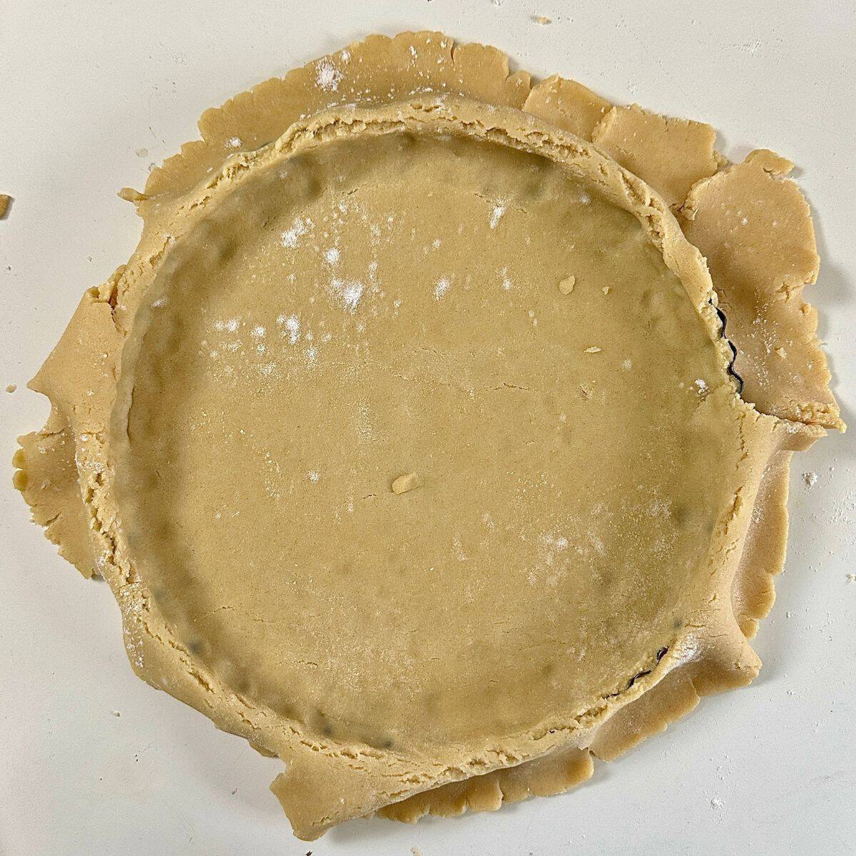 Pastry dough for a vegan quince frangipane tart pressed into a tart tin, with dough hanging over the sides, ready to be trimmed.