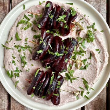 A white plate spread with a layer of bortolli bean dip with three gochujang beetroot skewers placed on top.