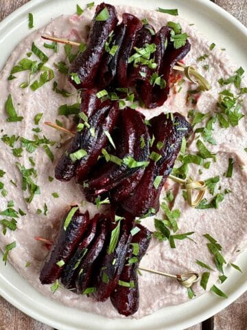 A white plate spread with a layer of bortolli bean dip with three gochujang beetroot skewers placed on top.