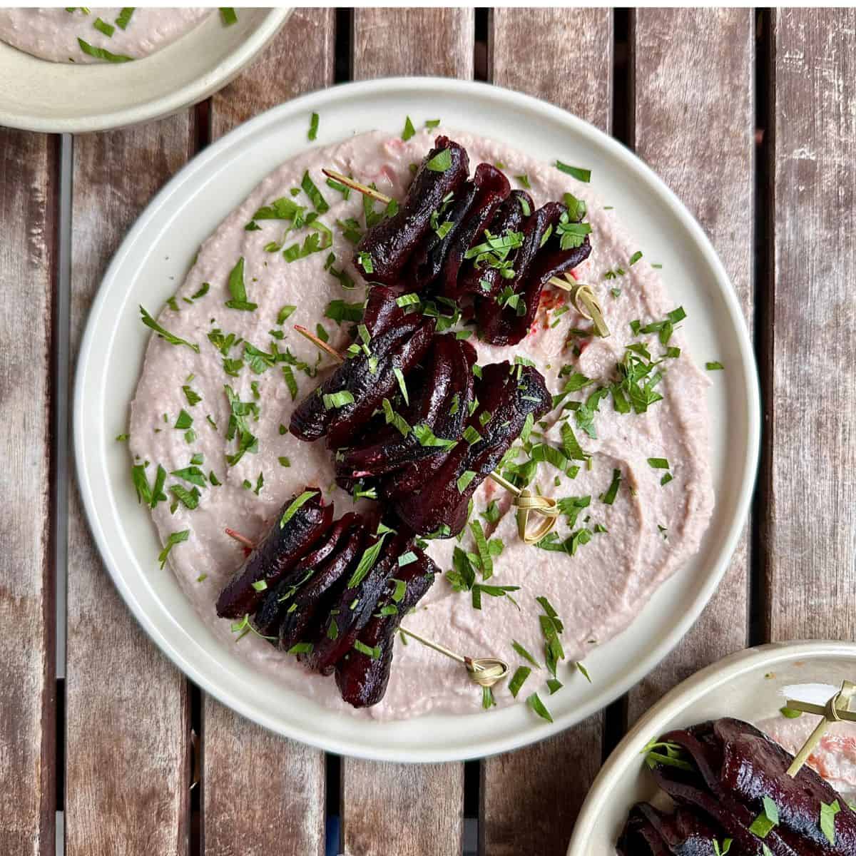 A white plate containing a bed of borlotti bean dip and three beetroot skewers sitting on the borlotti bean dip.