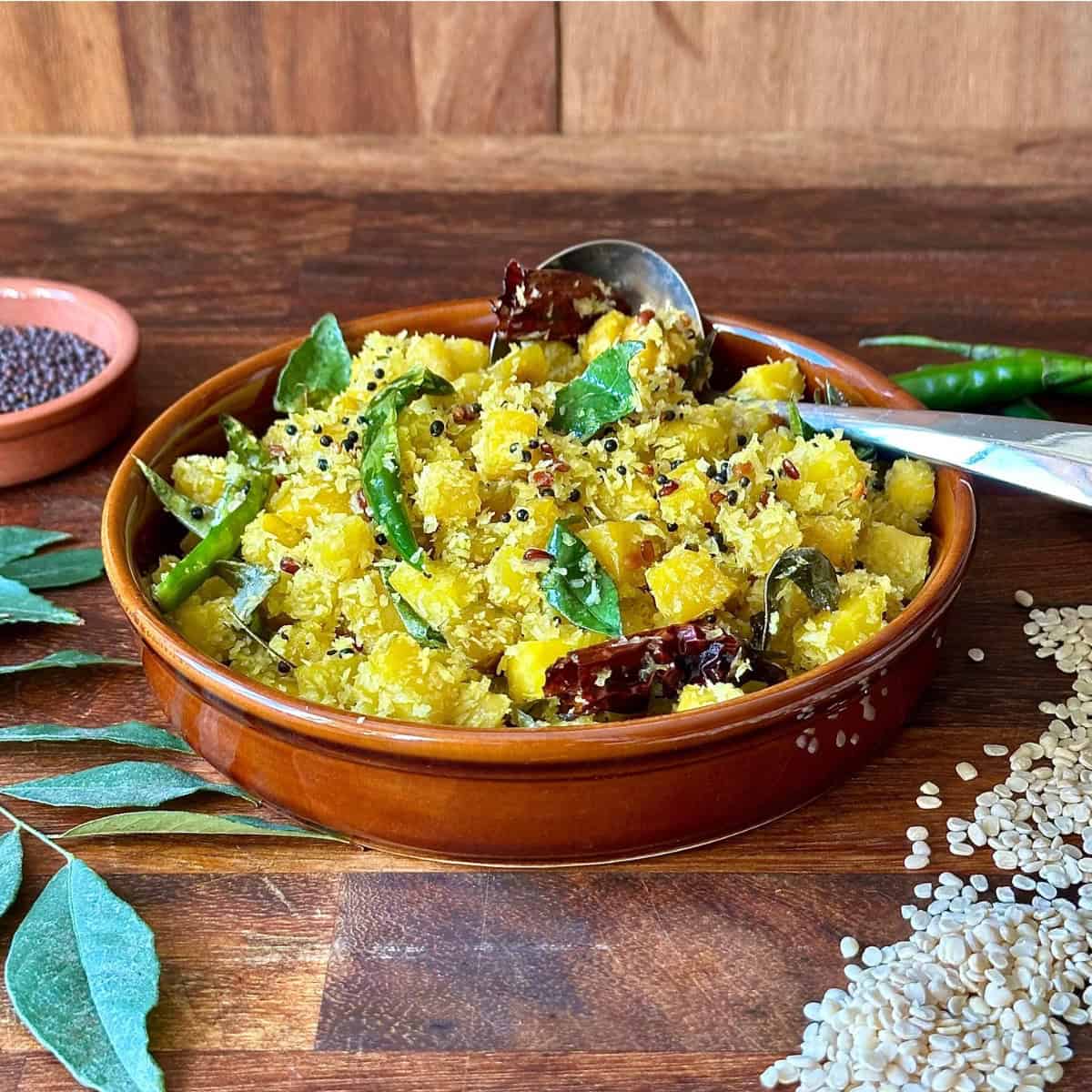 A shallow brown dish containing golden beetroot thoran, with a spoon rising in the thoran. a stem of curry leaves, some uncooked urad dal and a dish of black mustard seeds surround the thoran as decoration.