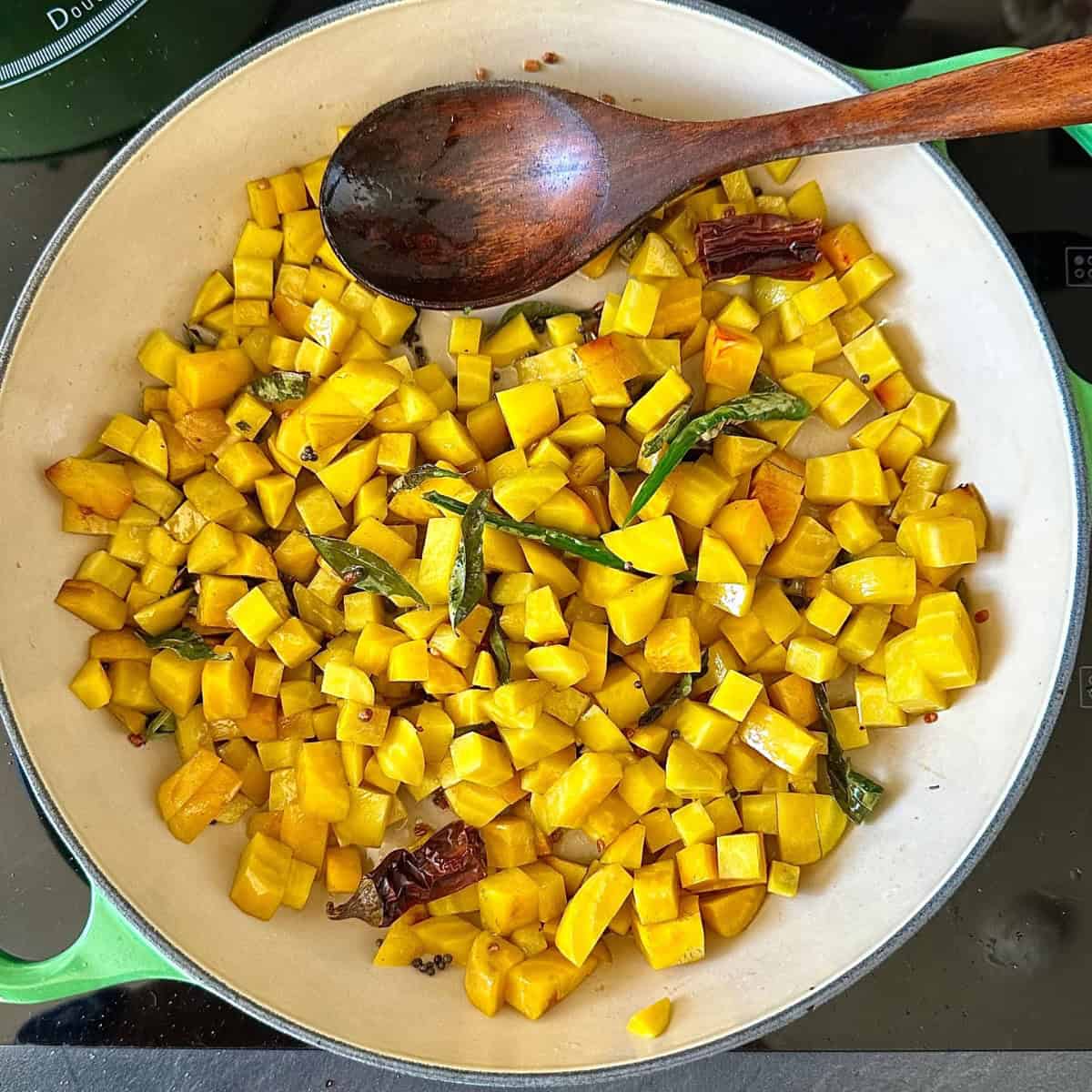 A large green casserole contains golden beetroot dice cooking with a black mustard seed, curry leaf, dried red chilli and green rocket chilli temper, for golden beetroot thoran recipe.