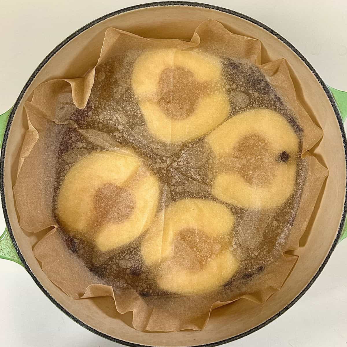 A large green pot with 4 peeled cored and halved poached quince, covered with a parchment paper cartouche. For a quince frangipane tart recipe.
