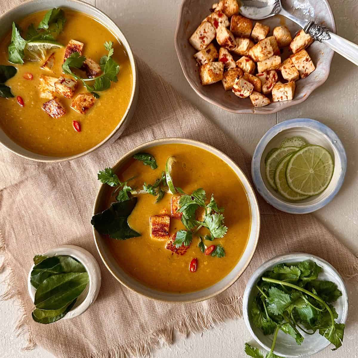 Two bowls of thai pumpkin soup with smoked tofu cubes and garnished with coriander. Surrounded with bowls of lime leaves, lime slices, coriander and cubed smoked tofu.