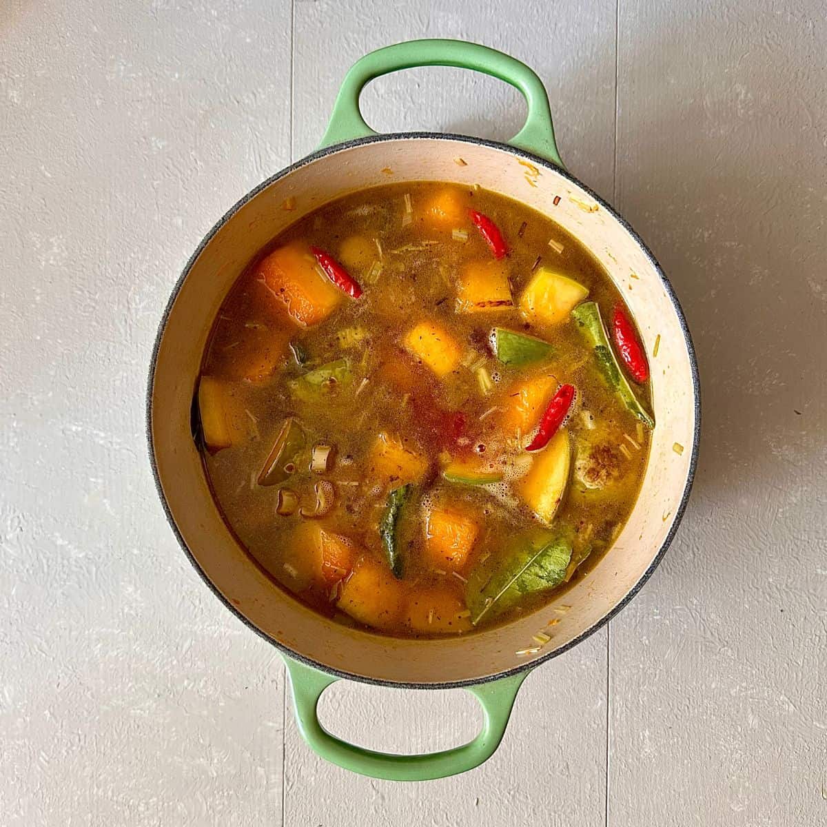 A large green pot containing diced pumpkin, lemongrass, bird's eye chilli, lime leaves and vegetable stock after cooking, for a thai pumpkin soup.