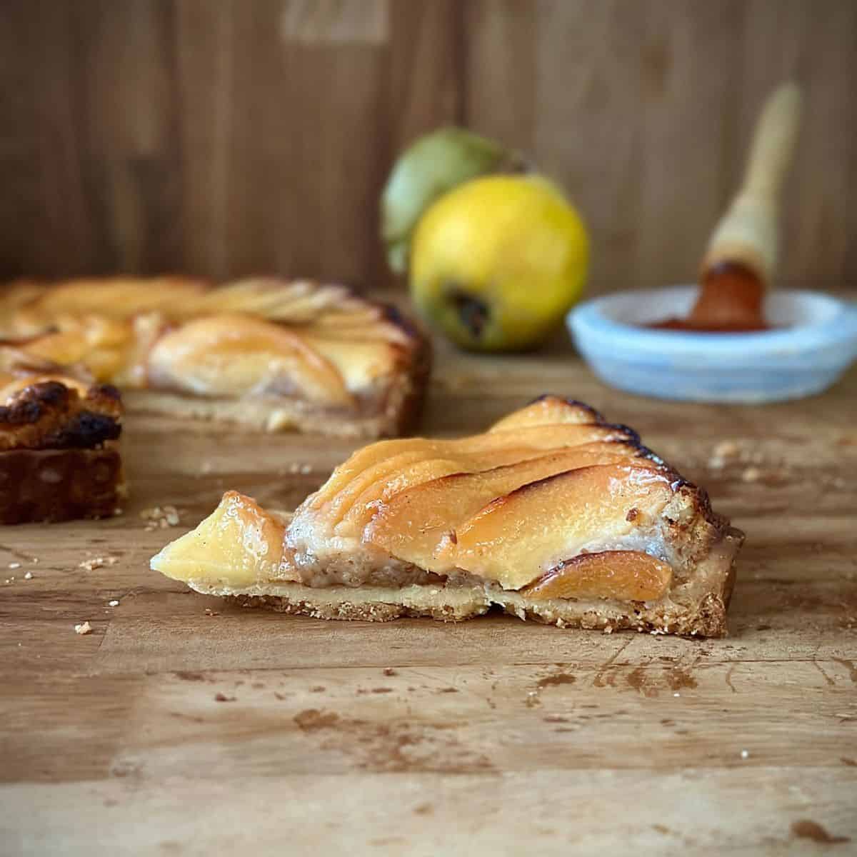 A slice of vegan quince frangipane tart sitting on a wooden chopping board. The rest of the tart sits in the background.