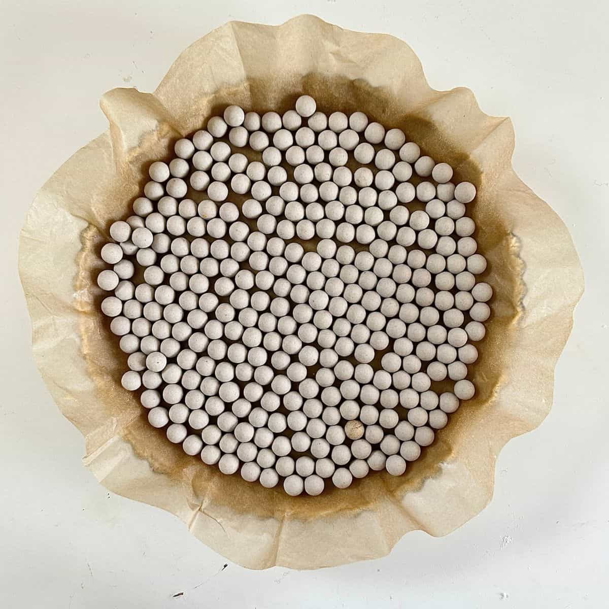 A vegan quince frangipane tart pastry base in a tart tin. The pastry is covered with parchment paper and ceramic baking balls have been placed onto the parchment paper, ready for baking.